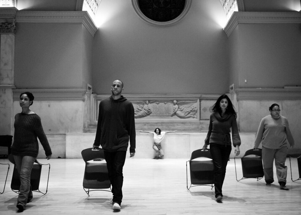 A photo of dancers performing at the Judson Church. Four dancers walk towards the camera dragging chairs behind them. At the center and in the background Marya Wethers supports her body with outstretched arms at the altar. Photo by Ian Douglas