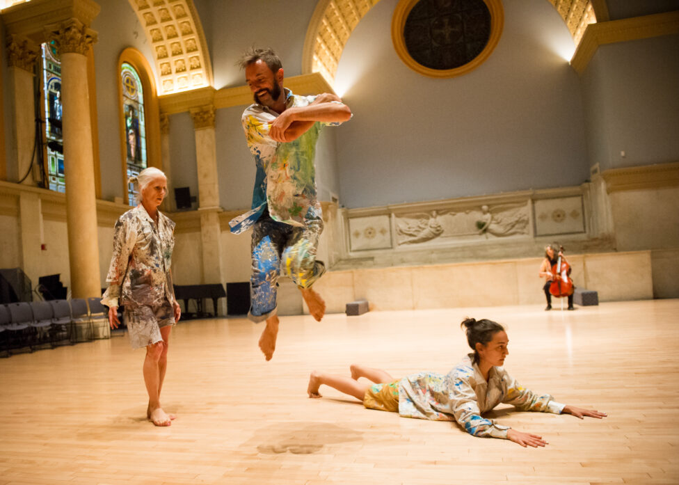 A photo from K.J. Holmes' performance for Movement Research at the Judson Church. Three dancers int he foreground. K.J. stands while a second dancer is caught in mid air smiling. A third dancer lays on the floor on their stomach with their torse propped up by their forearms. They all wear paint splattered clothing. In the background a musician plays the Cello. Photo by Ian Douglas.