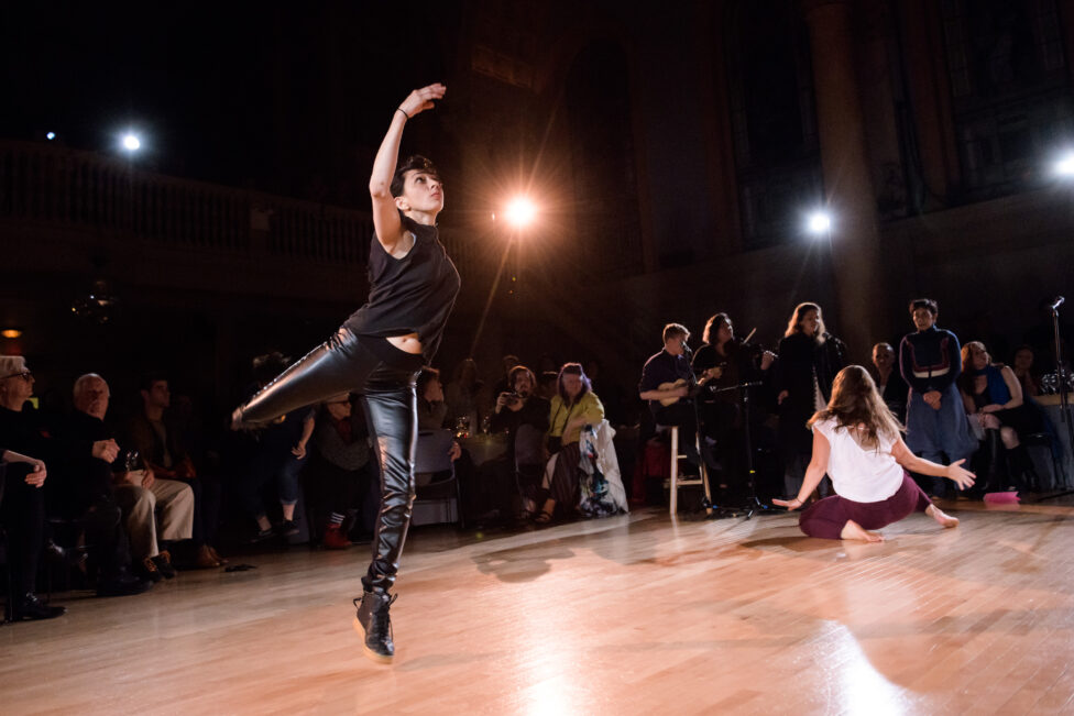A photo from the 2028 MR Gala. One dancer steps forward with their other leg in a attitude position and one arm reaching up. Another dancers moves close to the ground. There are musicians and a singer. An audience watches all around them. Photo by Ian Douglas.