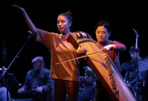 Kim and Clae (left and right respectively) are lit by a warm light as they guide audience members through a breath and movement meditation. Kim has her right arm stretched up while Clae strums their 古筝 with a bow. Photo by Kevin Yatarola.