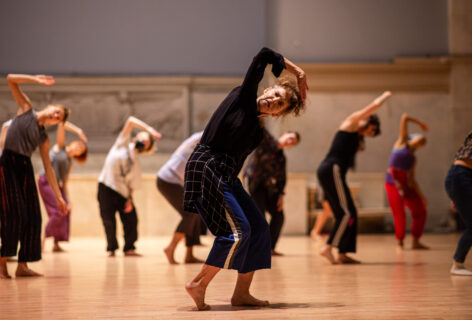 Photo of Vicky Shick teaching for Summer MELT at the Judson Church. She stands in front of the class while the dancers follow her. Her body weight is shifted on one leg while both her legs are bent. She is in a slightly crouched position, her torso curves to one side and her arm comes up with a bend at the elbow. Photo by Rachel Keane.