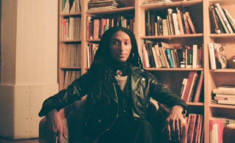 Ke'ron sits cross-legged in a chair in front of a large bookshelf. She has long black twists for hair, sports silver hoop earrings, and is wearing a leather jacket that covers her black vest and grey sweater. Photo by Grace Rosario Perkins.