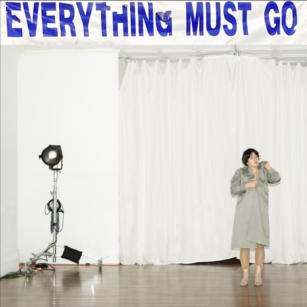 Tess Dworman wears a grey trench coat with beige tights and dance shoes. She stands in a room alongside a light and beneath a peeling sign that reads 