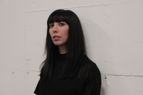 Roya Amirsoleymani, a brown, femme person, leans against a gray, concrete wall in the warehouse of Portland Institute for Contemporary Art. She has long, black hair, black bangs, hazel eyes, and a gold nose ring. She is wearing an all-black, short-sleeved shirt with a high neckline. The picture is cropped at her midsection. Photo credit to Mia O'Connor.