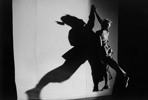 Photo of a dancer leaping and placing her hand on a wall. The dancer creates a large shadow on the wall and the image is in black and white. Photo by Green Deng.