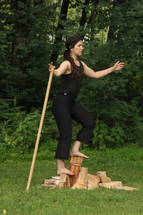 Rori stands barefoot on a pile of chopped wood in front of leafy trees. Her arms are outstretched and she drags a wooden staff slightly behind her. She looks far away.