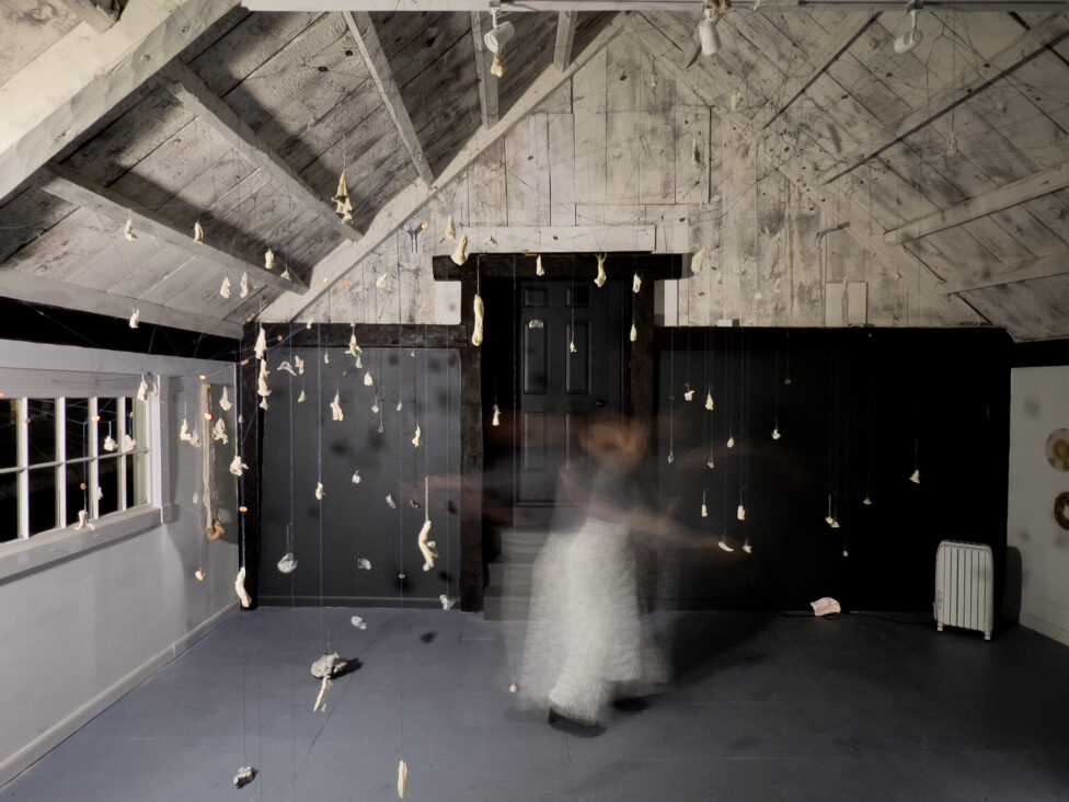A long exposure photo of Rori Smith moving in a room filled with objects dangling on cording, hanging from the ceiling. 