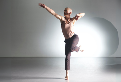 An action shot of Jennifer Nugent in a bright space. A light spotlights behind her as she holds her arm out and balances on one foot. She wears a pink top and grey striped pants. Photo by Jim Coleman.