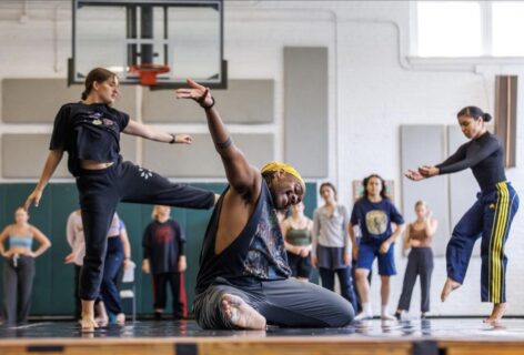 A tall black dancer is on the floor with his right arm extended on the high diagonal. His right leg is extended and reaching toward the camera. He is foregrounding a room full of people in a large dance space with a black floor. Photo by Ben McKeown.