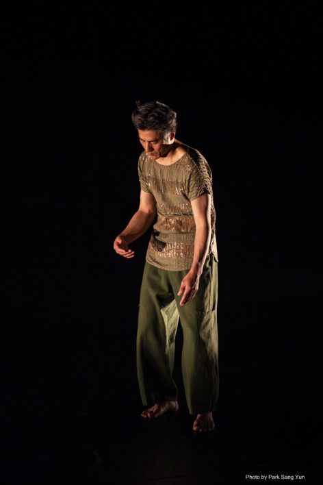 Medium-build Asian male with dark, mid-length hair, facing diagonally to his right with face downcast; wearing light-green top with gold accents and forest green pants. Dark background. Photo by Park Sang Yun.