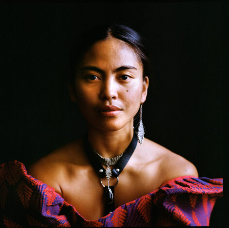A brown, Filipina woman looks straight forward towards you, with a grounded, kind, haunting expression. Her hair is black, slicked back and parted down the middle. She wears one silver chain earring, matching her choker necklace with black obsidian for protection. She wears a black leather harness underneath a red and purple traditional Filipiniana terno, made by traditional weavers near her mom’s home in Batangas. The terno butterfly sleeves are uneven, the one on her left shoulder has fallen to the side. Photo by Maria Baranova.