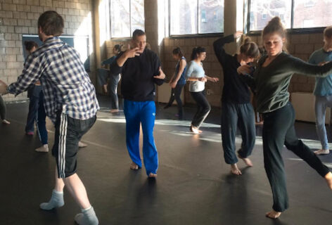A dance studio filled with dancers wearing casual dance clothes, each in the midst of different actions and shapes. Neil, a white cis-man wearing loose sweat pants and a longsleeved shits, is caught mid-step with hands in front of him in gesticulation. Photo courtesy of the artist