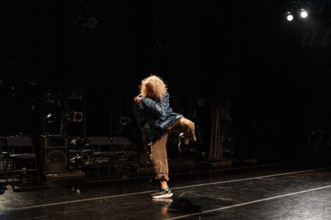 Sara Shelton Mann in performance. She stands on black marley wearing brown cut pants and a blue jacket. She is balancing on one foot with one arm hugging her stomach and the other in front. Photo by Robbie Sweeney
