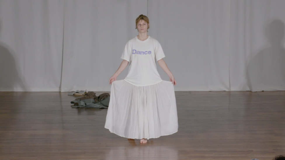 With a dissociated expression, Sonya Gadet-Molansky lifts the two sides of their skirt during a moment in Everything Must Go. They wear a white t-shirt with 