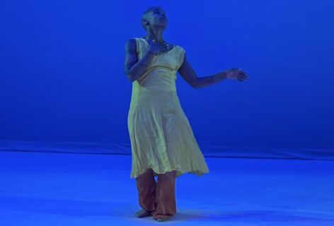 A blue lit stage with Jasmine Hearn in a yellow dress and orange pants. They look up with their hand to their chest one arm bent and out. Photo courtesy of artist.