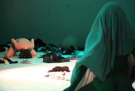 A photo of luciana in performance of PUROTEATRO. She is bent over with her body uncovered and her head resting on the floor. there are piles of clothing all around and a green wall behind her. In the foreground there is a large pile with a green fabric draped over it. Photo by Brian Rogers.