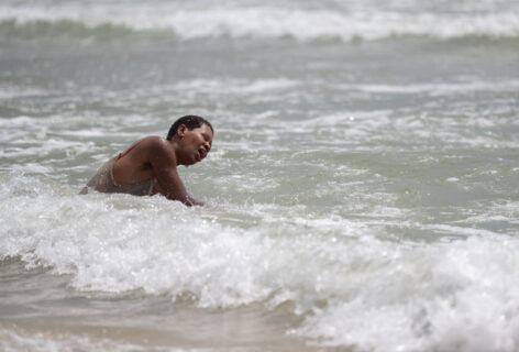 Brown skinned person with short black kinky hair, moving sideways in the ocean, engulfed by waves. Photo by Nir Arieli