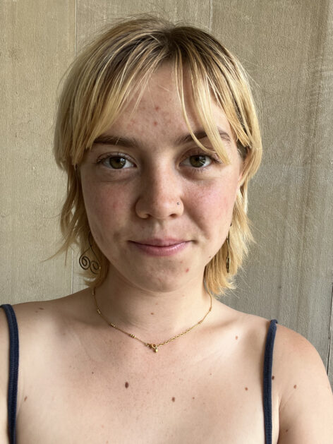 ID: Close up portrait of a white woman in a tank top with short bleached blond hair smiling at a camera.