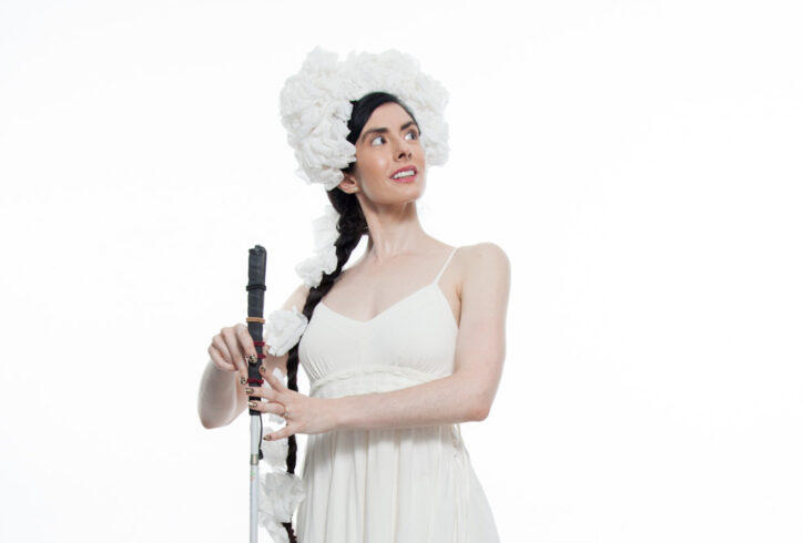 Krishna, a thin, pale skinned woman with long dark hair pulled into a braid, poses on a studio stage. She is wearing a simple white dress with her shoulders exposed and a fluffy white headdress perched on her forehead and wrapping down her braid. She looks over her left shoulder as she lightly holds a straight cane between her hands and points her left foot behind her. Photo Credit: Krishna Washburn & Micaela Mamede