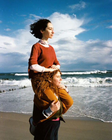 Image of Sophie sitting on Lindsey's shoulders at the Beach. Sophie has short dark hair and wears a red t-shirt over a white Turtle neck and orange pants. Lindsey has ginger hair and wears a dark green cardigan and black pants. They both face the sun with windswept hair. Photo By Ivy Meissner.