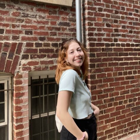 ID: Ruby stands in front of a brick wall, smiling over her right shoulder to the camera.
