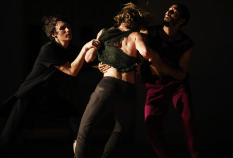 Three male dancers in a trio situation fraught with tension, in a dark theater space. On person shirt is being pulled and all are looking in different directions. Photo by Paula Court.