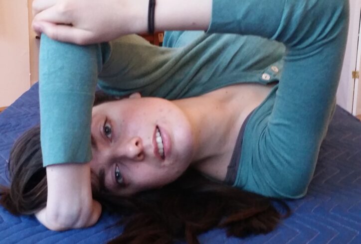 Anat lies on a blue quilted mat. She wears a long sleeve teat top. She looks into the camera, one arm reaches over her head, palm pressed into the mat. The other reaches across to catch the elbow and stabilize. Photo courtesy of the artist.