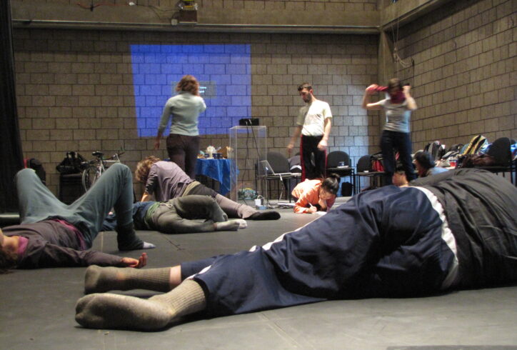 From the MR Archives. Moving Dialogue, Romania, 2010. Photo of participants in various states of rest and observing in a room with brick walls. A blue light is projected in the background.