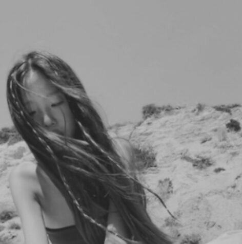Grey tinted photo of Autumn, sitting by a cliffside, gazing down towards the sand (unpictured). She is wearing a bandeau as the wind sweeps her hair to the right side of the frame.