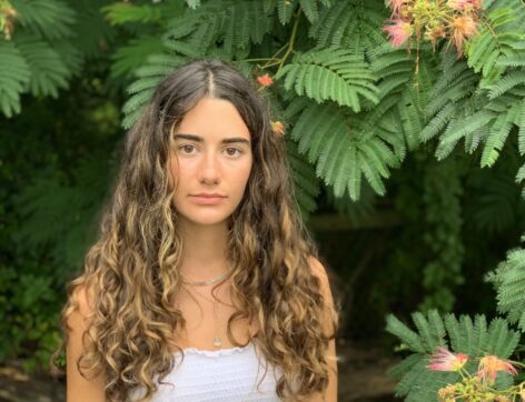 ID: Portrait of Grace from the shoulders up standing in front of a Mimosa tree. She is smiling slightly and has long brown curly hair, brown eyes, and is wearing a white tank top. By Katherine Hammond.