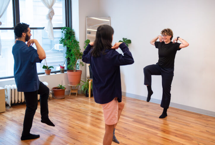 Kari Ostensen teaches a class. Kari faces the students demonstrating a position they attempt to replicate it. They all balance on one leg while the second leg is bent at 90 degrees in front of them. Their arms are bent at the elbows and fingertips touch their shoulders, their elbows reach to their sides. Photo by Didi Zacks.