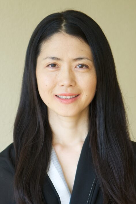 The image features a close-up headshot of a person with long, straight black hair. They are smiling softly and looking directly at the camera, giving a warm and approachable impression. Their skin is clear, and they have minimal makeup, with a natural look. They are wearing a black garment with a white inner lining, suggesting formal or traditional attire. The background is a plain, neutral color, which keeps the focus on their face. Photo by Love Hellgren