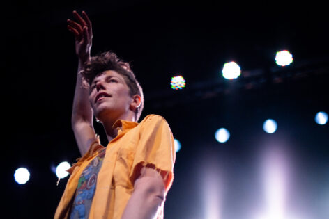 Shot from below, looking up - a white woman with short curly hair, wearing yellow and grey shirts, stands with her right arm extended above her head, lookin out. Theater lights are visible behind her. Photo credit to Anna Maynard.