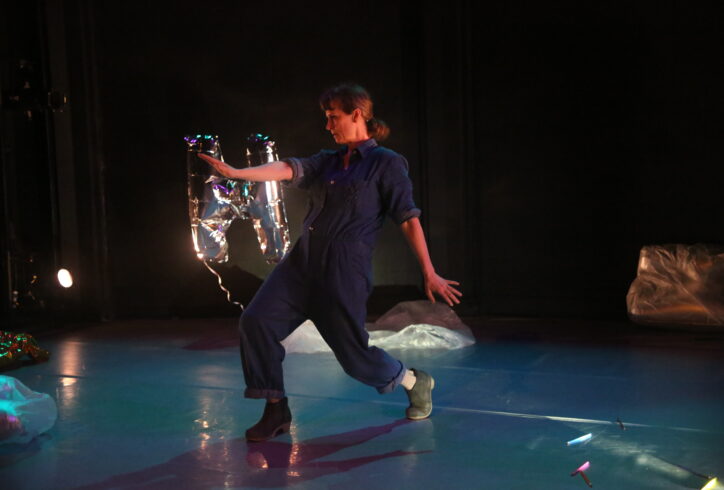 A white woman in a blue jumpsuit is hinging back into a lunge with one arm forward and splaying her other hand back. There is a a silver helium 
