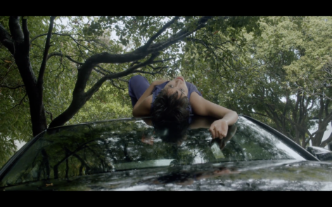 ID: Andrea's body lies on top of a black car, with her head and arms down the front window. She is wearing a purple shirt, and red lipstick. Green trees in the background.