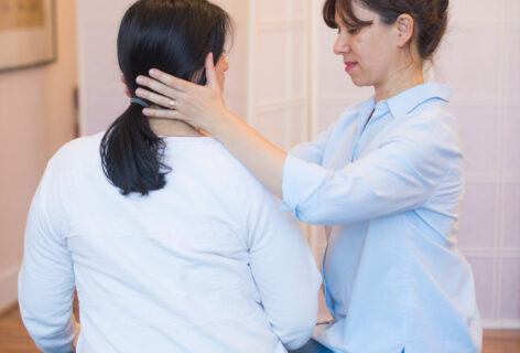 A graphic with text over a photo. Anat is in session with a client. She sits next to the client and places her hands at either side of the clients lower jaw and neck. Photo by Elodie Saracco