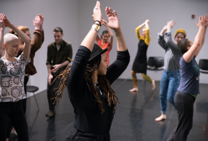 A group of dancers in a black box studio dance in a circle. Photo by Asya Gorovitz