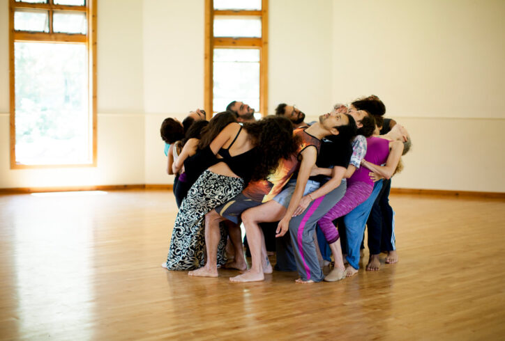 A group of dancers stand in a circle. They all squat and lean their torsos on each other creating a supportive spiral. Photo by Anna Maynard.