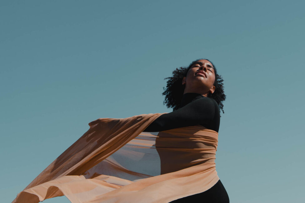 An image of Megan Curet in movement. Below a clear blue ski, Megan tilts her face towards the sun, eyes closed and coily hair floating in the wind. She wears a black turtle neck and moves a sheer brown fabric around her torso with her arms. Photo courtesy of the artist.