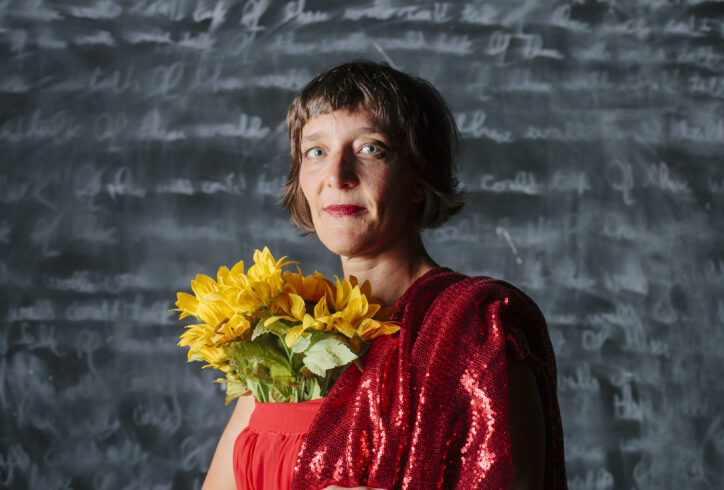 A close-up portrait of Esther Baker-Tarpaga wearing a red sparkly dress with a bouquet of yellow flowers poking out of the top. She is smiling softly at the camera. Behind her is a chalkboard with blurred lettering. Photo by Karla Conrad.