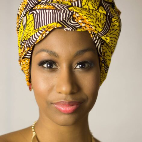 Milteri Tucker, a person with brown skin and bright eyes, smiles to the camera while wearing a vibrant yellow headscarf.