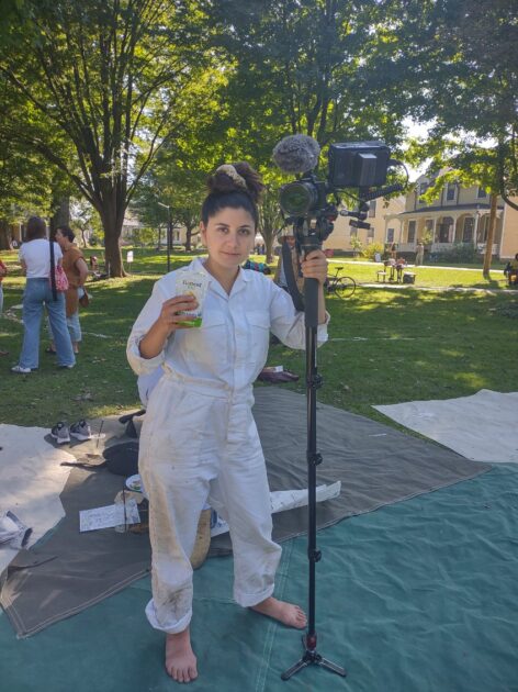 White woman in a khaki jumpsuit grins softly while holding film equipment and a juicebox under the trees on Governor's Island