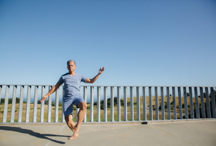 A brown-skinned Filipino man, Gerald wears a black t-shirt and jeans and dances against a concrete wall and Redwood trees. Photo by Crystal Birns.