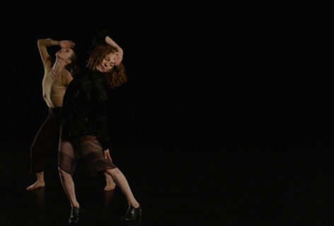 Jodi Melnick and another performer in motion. Their hands on their heads and leaning foward the left with their chest up over a black background. Image by Kate Hawkins.