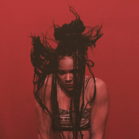 j alex ray (a tan person with braids) in front of a red background. Photo courtesy of the artist.