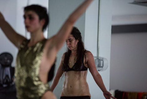 Two light skinned dancers' upper bodies one in front of the other. Front dancer is wearing a gold sequined leotard opened on the sides and back dancer is wearing a lace navy blue bra with pants pulled down to just above the pubic area. The dancer in front (Molly Lieber) has her arms stretched upwards in a V shape and the dancer in back (luciana achugar) is looking down and has her arms down by her sides also stretched out in a quasi V shape. Photo by Scott Shaw.