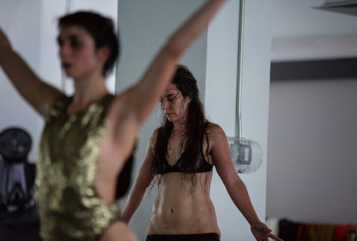 Two light skinned dancers' upper bodies one in front of the other. Front dancer is wearing a gold sequined leotard opened on the sides and back dancer is wearing a lace navy blue bra with pants pulled down to just above the pubic area. The dancer in front (Molly Lieber) has her arms stretched upwards in a V shape and the dancer in back (luciana achugar) is looking down and has her arms down by her sides also stretched out in a quasi V shape. Photo by Scott Shaw.