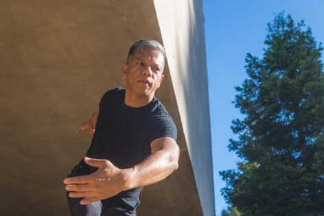 A brown-skinned Filipino man, Gerald wears a black t-shirt and jeans and dances against a concrete wall and Redwood trees. Photo by Crystal Birns.