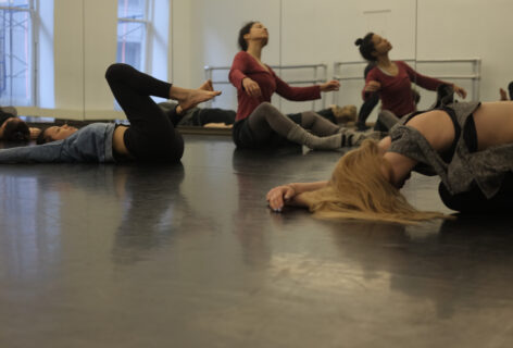 A group of dancers are on the floor in sitting and lying down positions, in the midst of moving. the photo is taken from a low angle. Photo by Lucy Sydel.