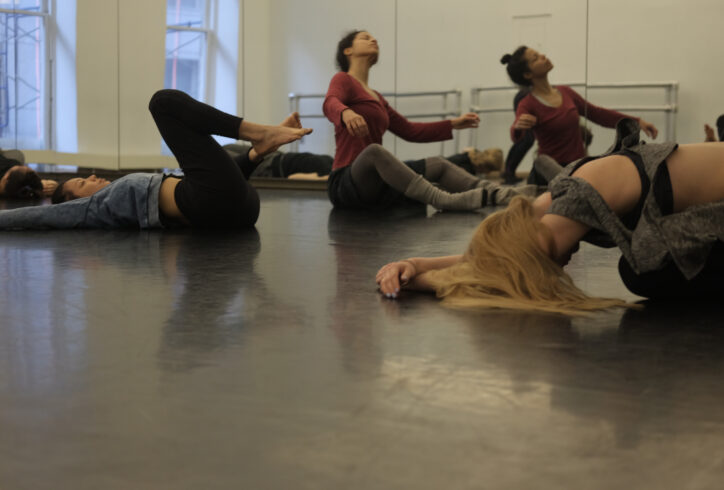 A group of dancers are on the floor in sitting and lying down positions, in the midst of moving. the photo is taken from a low angle. Photo by Lucy Sydel.